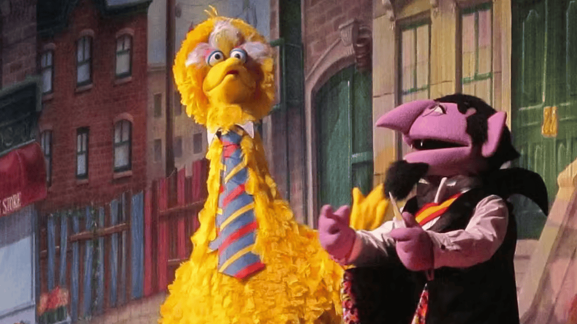 A colorful puppet of Big Bird and a puppet of a pink monster on a city street set.