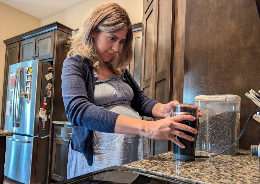 A woman stands at a granite kitchen counter. A refrigerator can be seen behind her. She is holding onto a coffee bean grinder and watching it grind coffee.