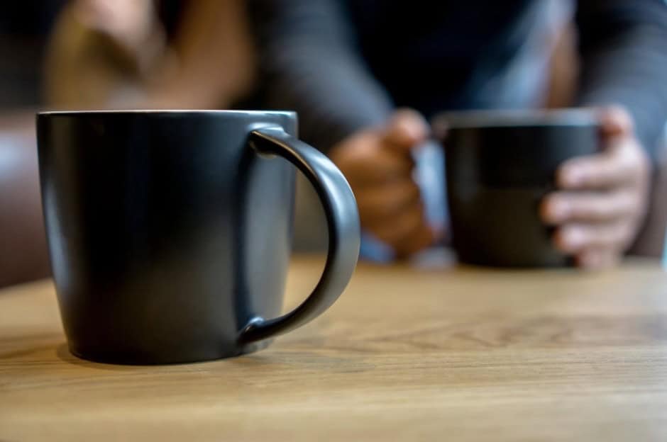 A black coffee mug on a wooden table with two people holding similar mugs in the background.