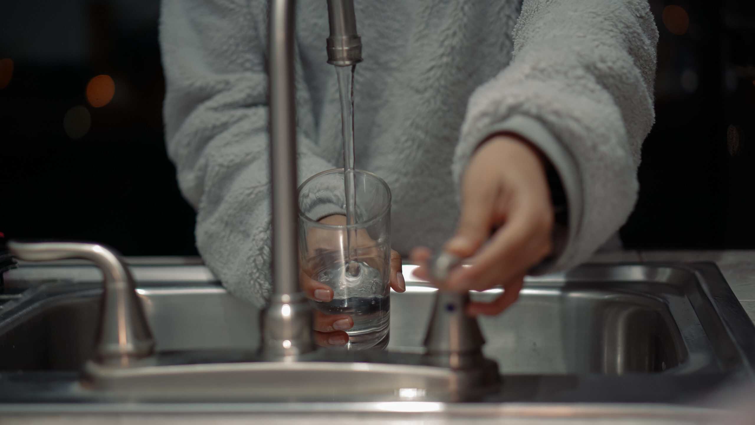 A person filling a glass with water from a kitchen sink faucet.