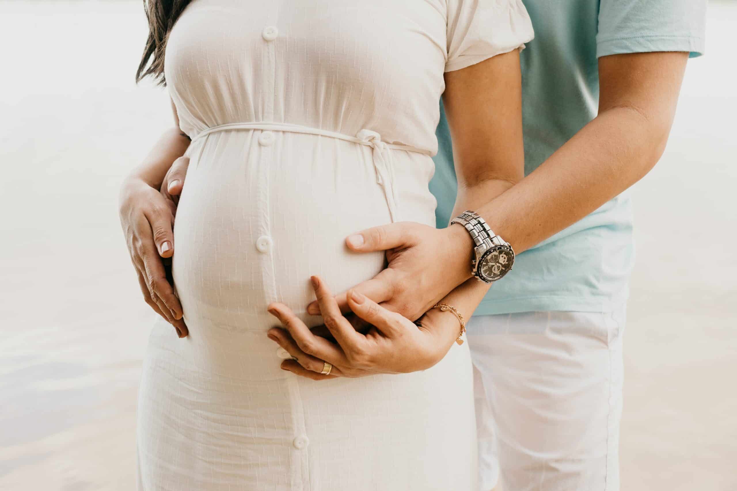 Pregnant woman with partner holding her belly at the beach, emphasizing support for Spanish-speaking.