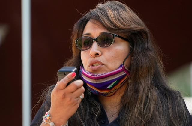Latina woman wearing sunglasses and a colorful mask, speaking into a walkie-talkie.