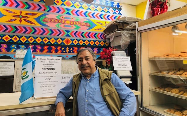 Fresh produce vendor smiling at Latin American market with colorful mural background.