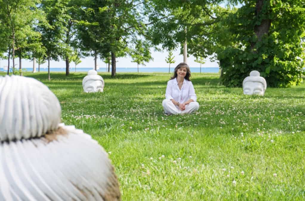 A woman practicing meditation outdoors in a lush green park with trees and sculptures.