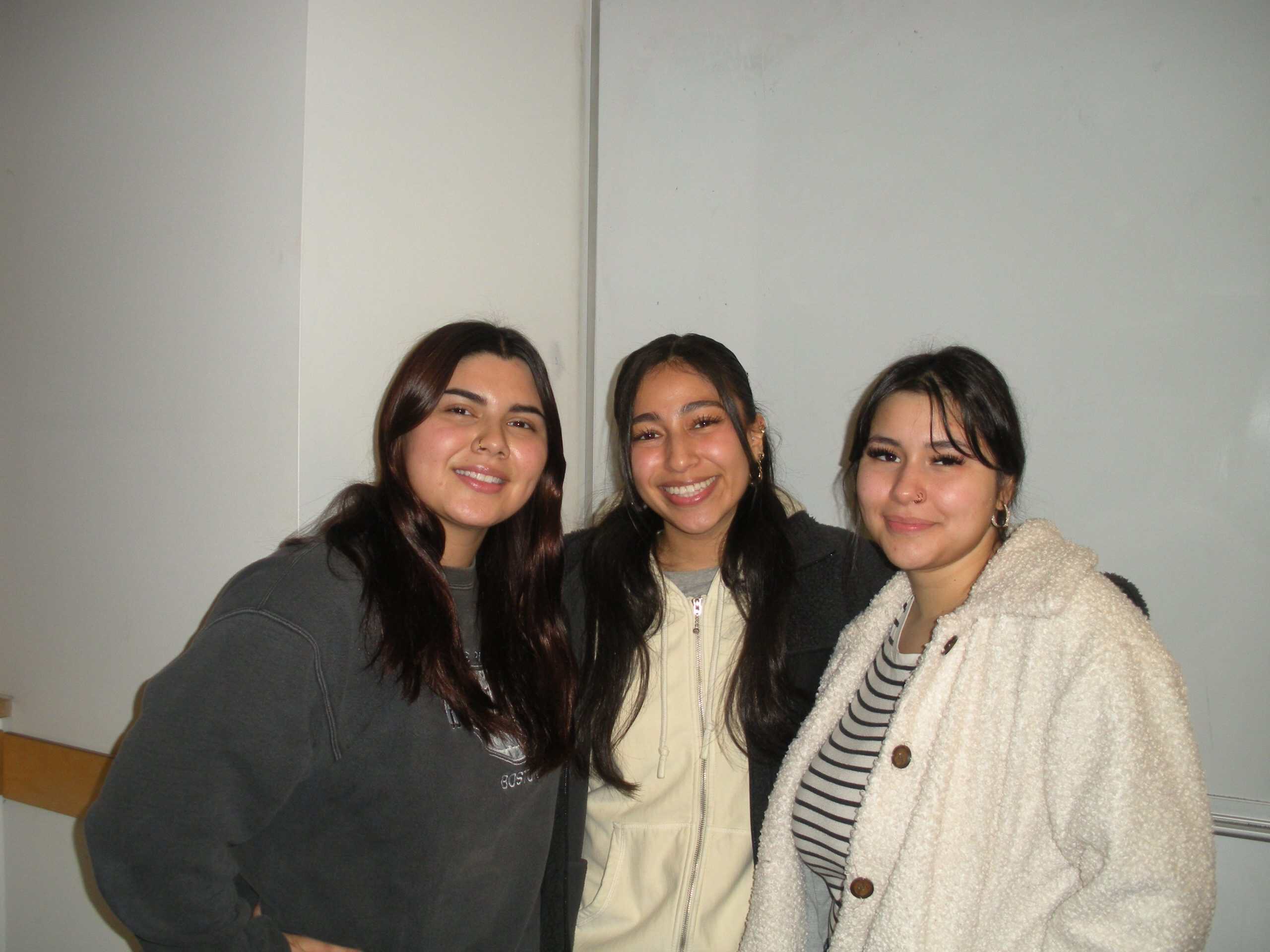 Group of three young women smiling indoors, casual attire, diverse backgrounds.