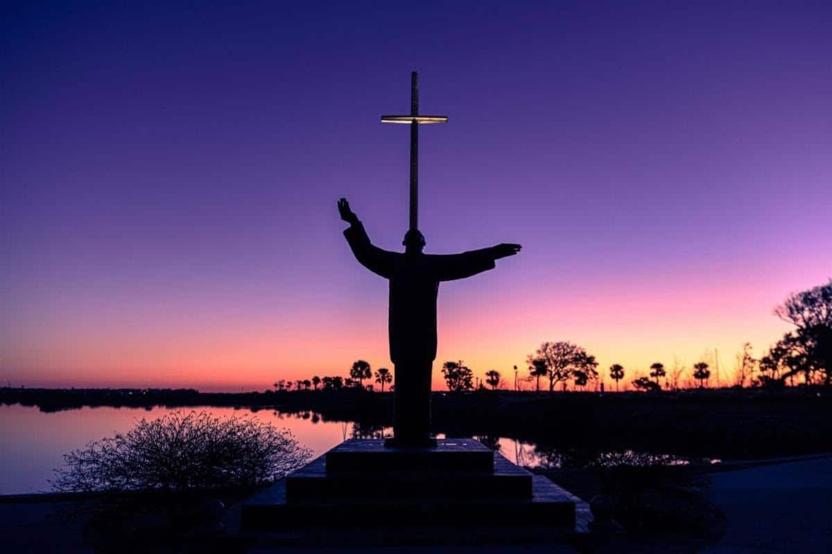 A person standing with arms outstretched in front of the Christ of the Redeemer statue at sunset in.