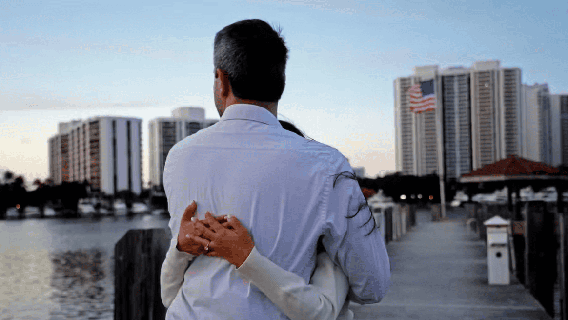 A woman hugging a man in a white shirt near a waterfront with city buildings and an American flag in.