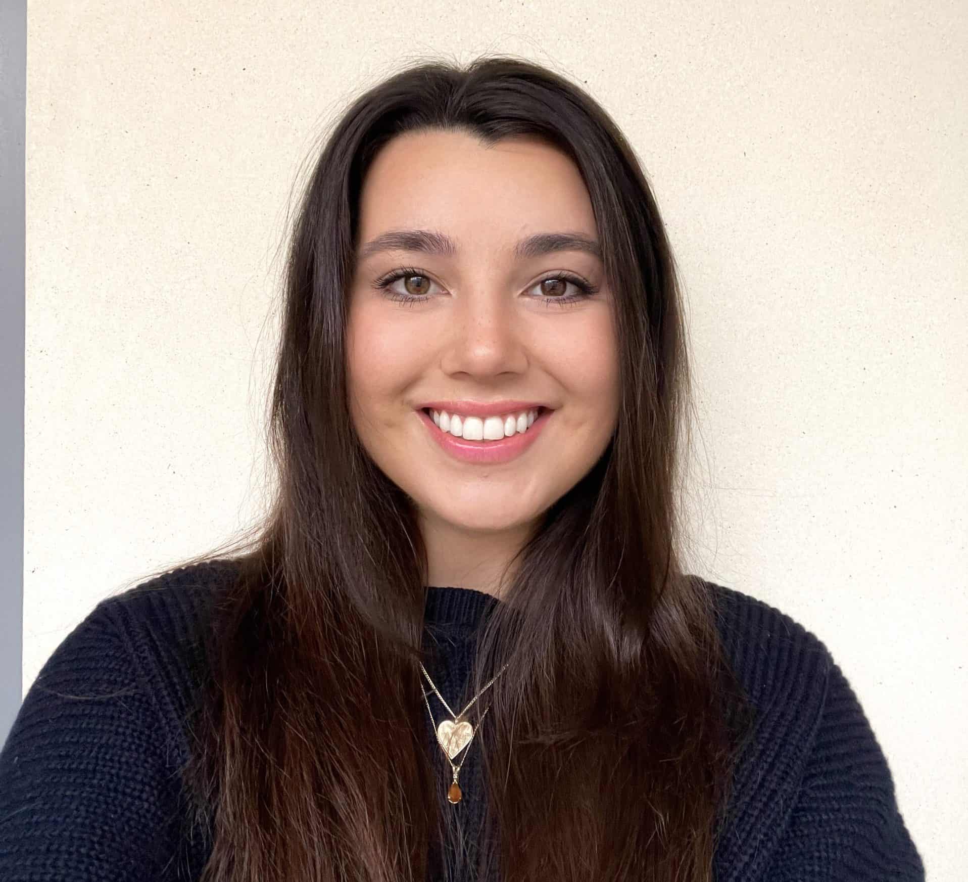 Bright young woman with long dark hair smiling, wearing a black sweater and a heart-shaped necklace.