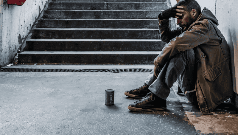 A homeless man sitting on stairs with a can in front of him, reflecting the need for housing as heal.